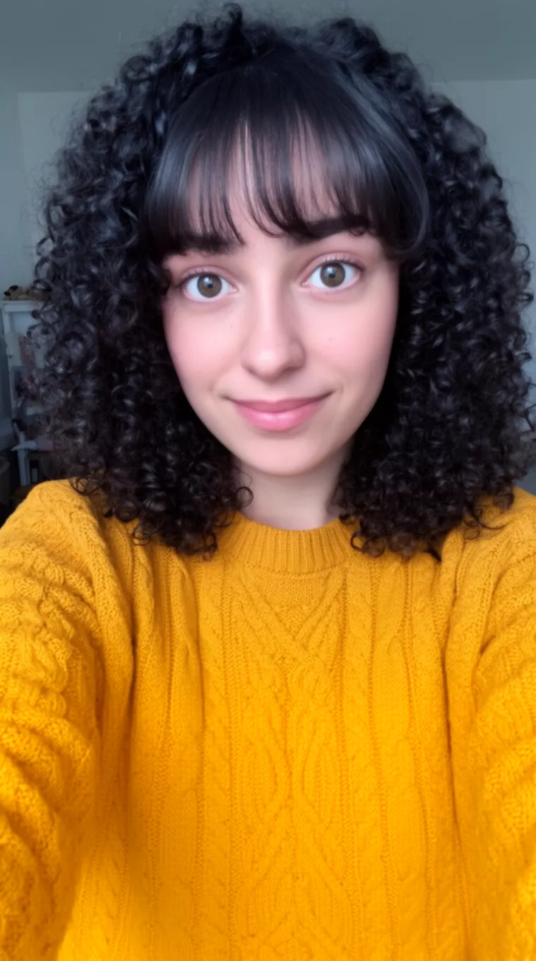 selfie of a 29-year-old woman wearing a mustard yellow cable knit sweater with a Curly hair with bottleneck bangs