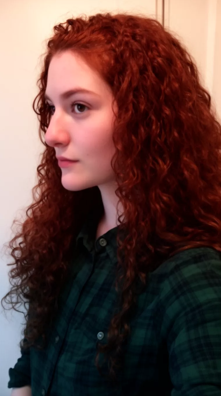 side view selfie of a 26-year-old woman wearing a dark green plaid shirt with a deep auburn brown long curly hairstyle