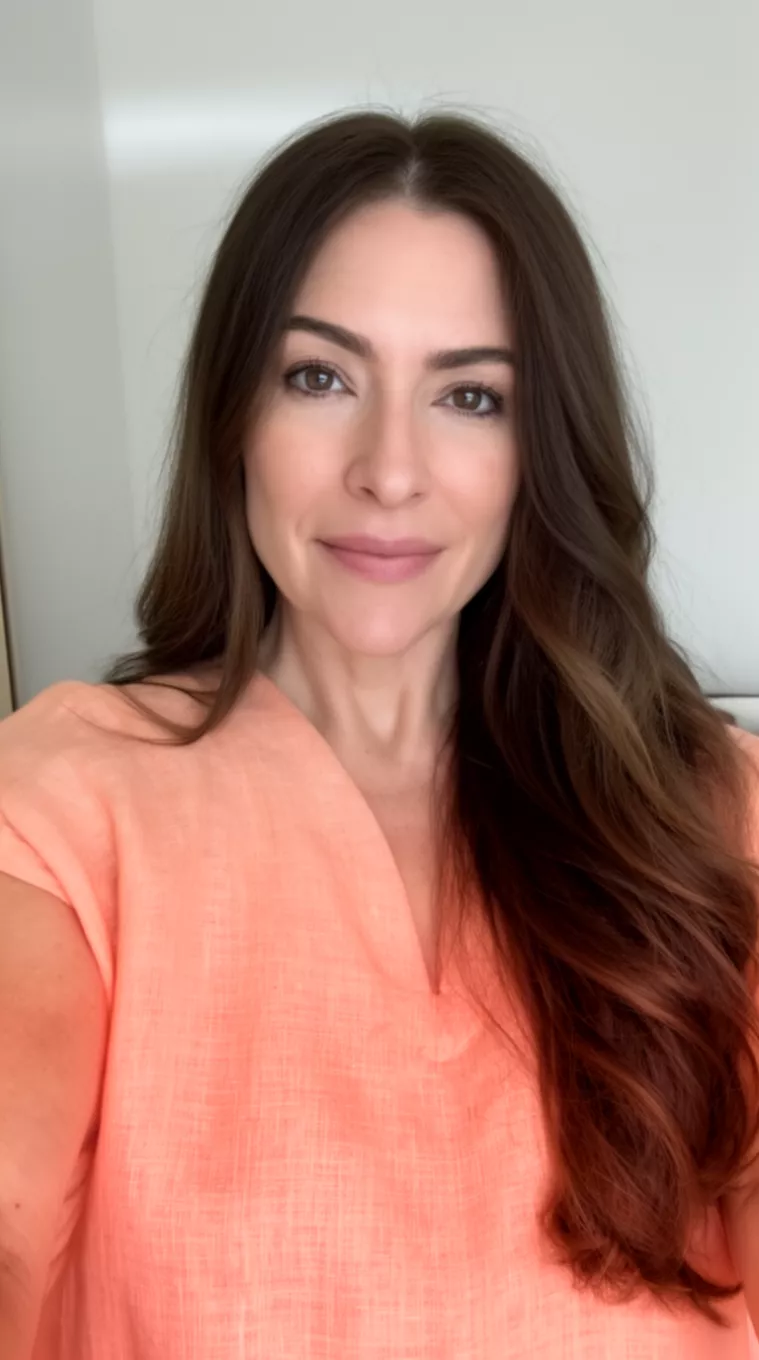 selfie of a 35-year-old woman wearing a peach linen top with a Hot Chocolate Brunette colored long hairstyle, minimal background