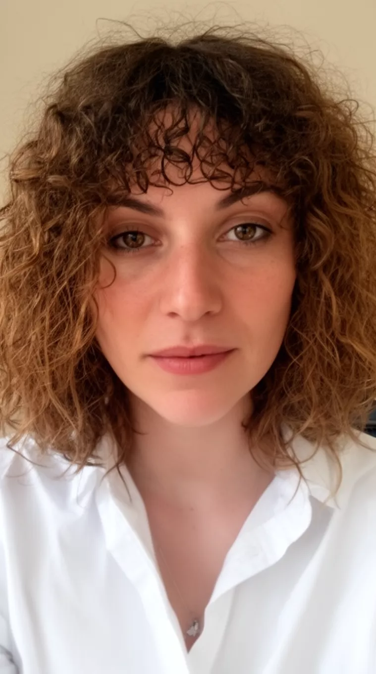 casual close-up photo of a 31-year-old woman wearing a white shirt with shoulder-length medium brown messy curly hair and curly bangs, minimal background