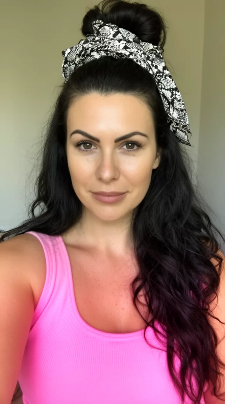selfie of a 36-year-old woman wearing a pink ribbed tank top with long dark wavy hair styled in a half-up high bun accented with a black and white snakeskin-patterned scarf, minimal background