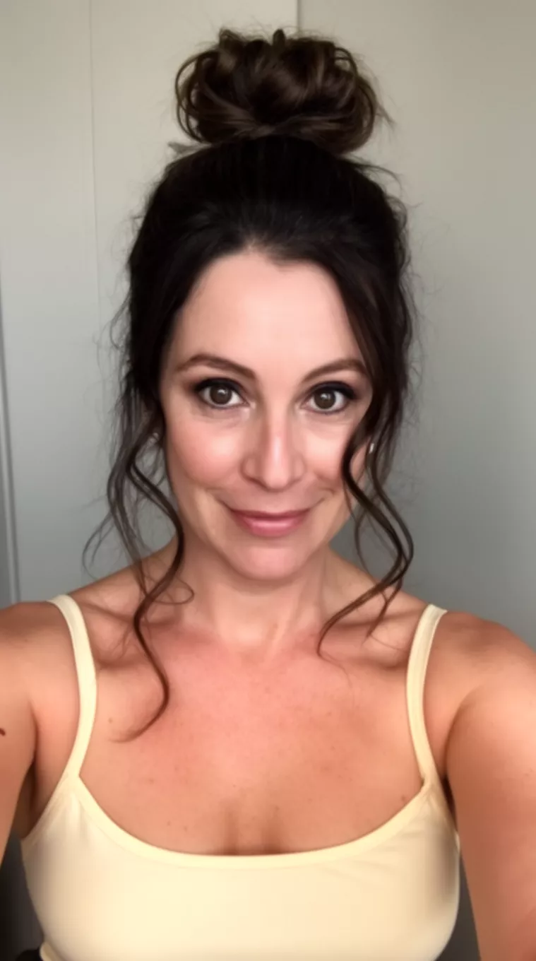 selfie of a 35-year-old woman wearing a cream-colored strap top with dark brown hair styled in a high messy bun with wavy tendrils curving near her face, minimal background