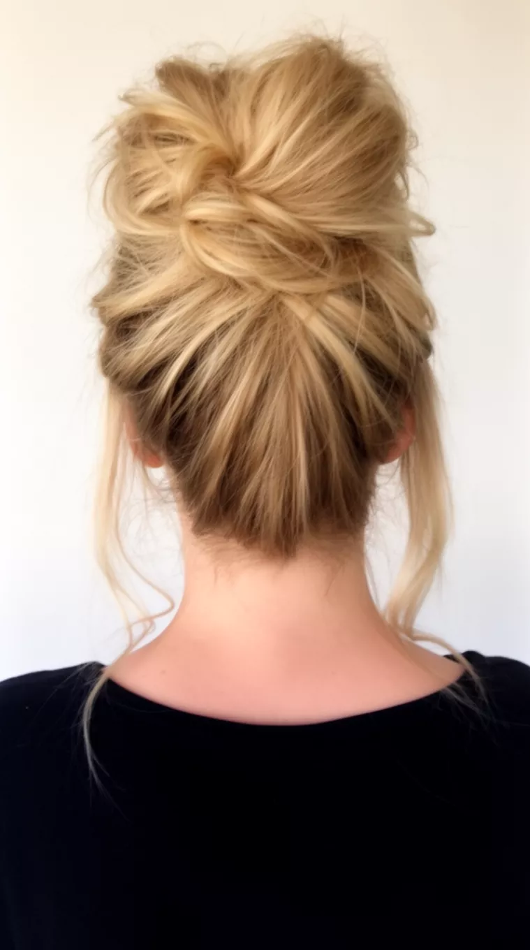 casual close-up photo of a 29-year-old woman wearing a black top with blonde hair styled in a voluminous messy high bun with face-framing pieces, back view, minimal background