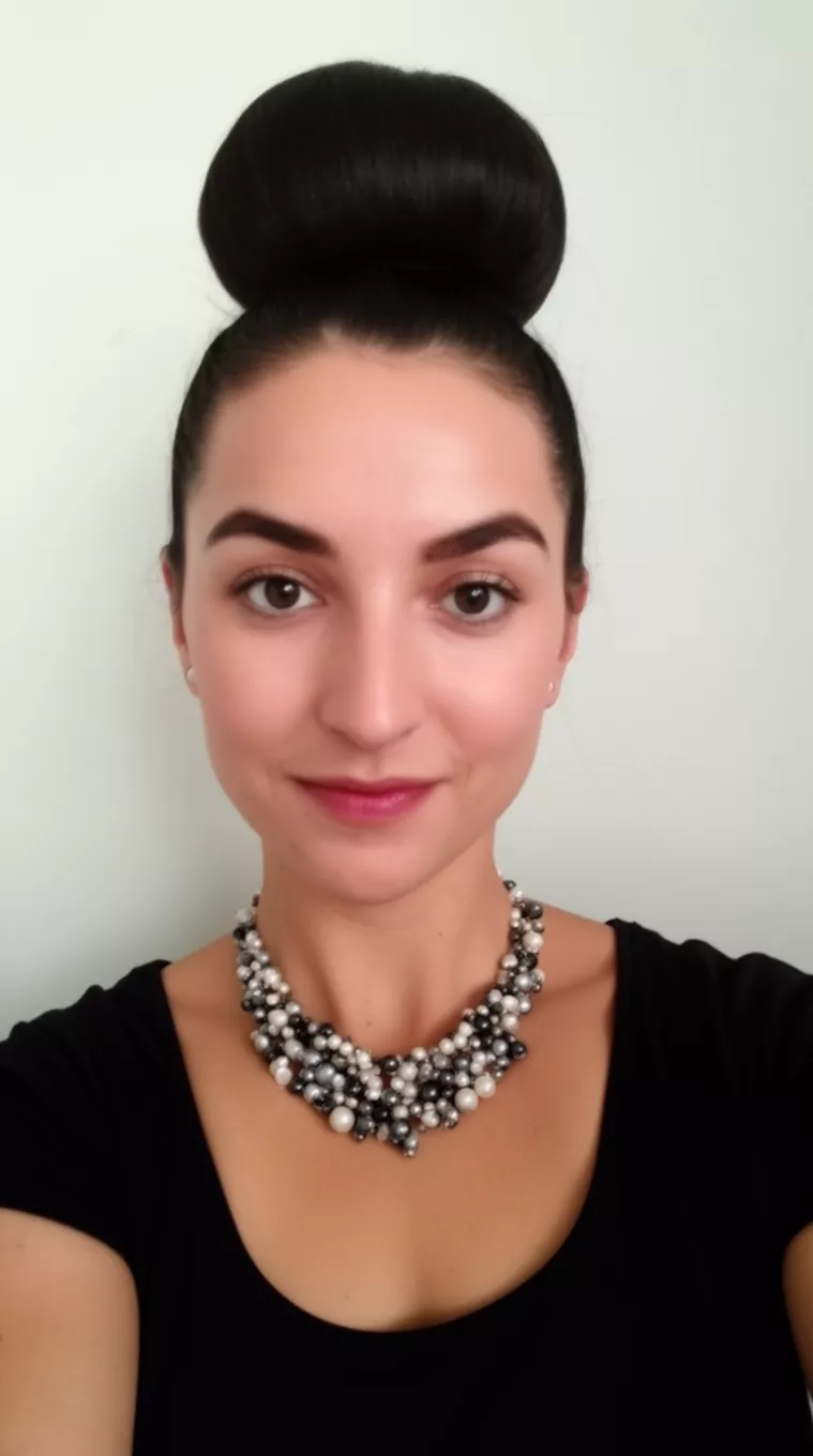 selfie of a 29-year-old woman wearing a beaded necklace with a very high, round bun hairstyle, minimal background