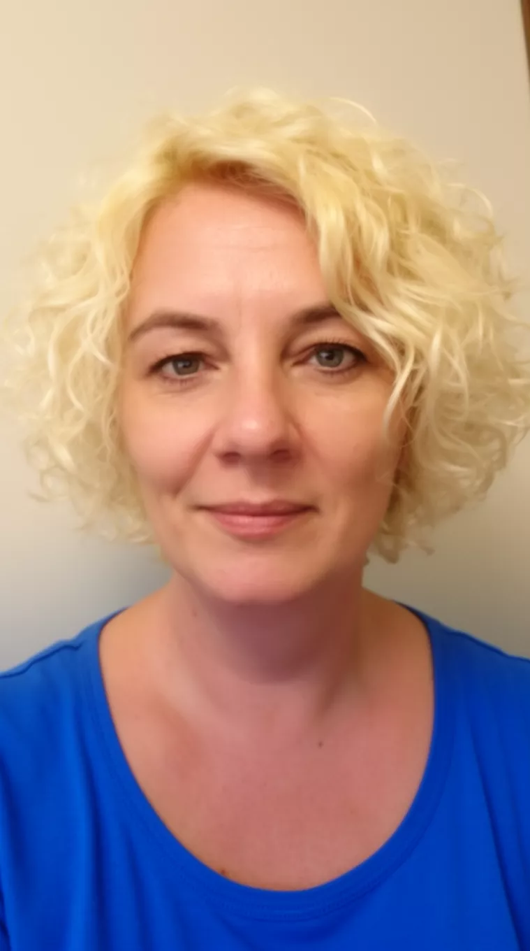 casual close-up photo of a 43-year-old woman wearing a blue top with short, wavy, bright blonde hair, minimal background