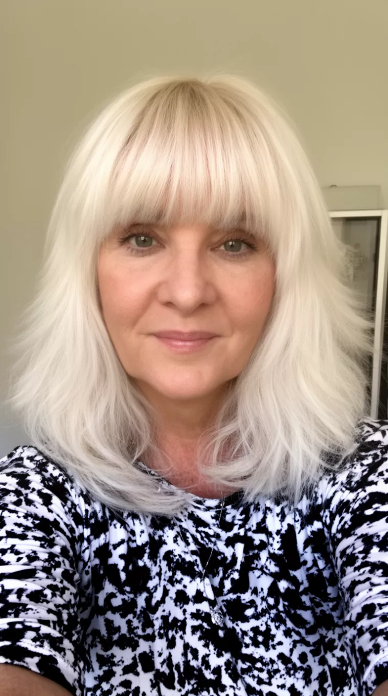 selfie of a 56-year-old woman wearing a black and white patterned top with a platinum blonde, layered medium-long hair with full bangs, minimal background