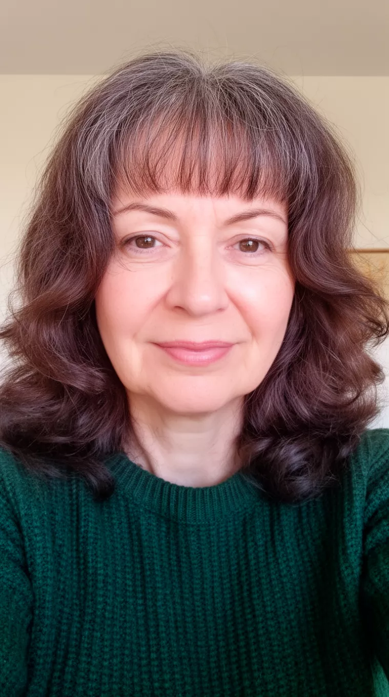 selfie of a 54-year-old woman wearing a dark green knit sweater with a mid-length, wavy, dark brown hair with subtle gray tones and short, wispy bangs, minimal background
