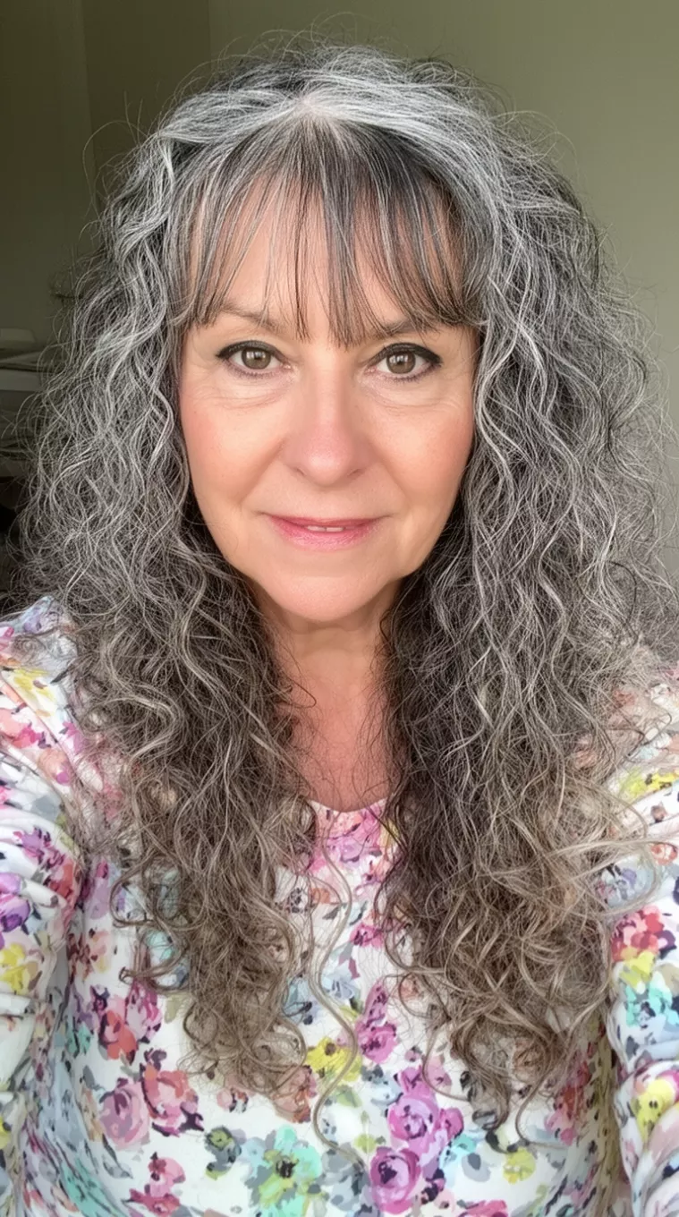 Salt and Pepper Curls selfie of a 64-year-old woman wearing a floral print top with long, curly, salt-and-pepper hair and fringe, minimal background