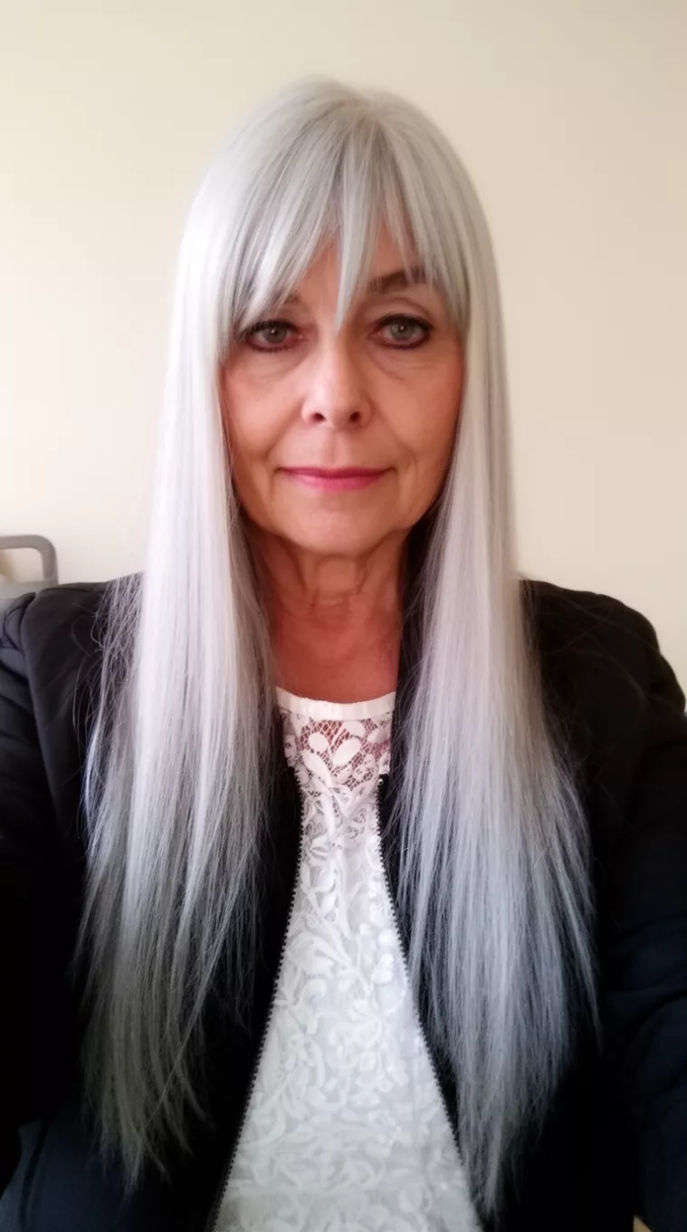 Long Silver with Side Bangs selfie of a 68-year-old woman wearing a black jacket and white lace top with long, straight silver hair and long side-swept bangs, minimal background