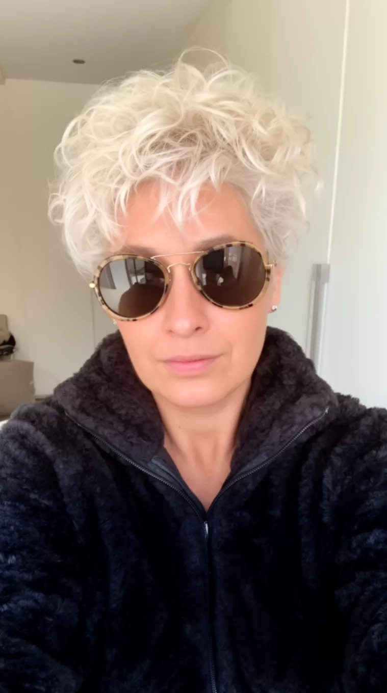 selfie of a 33-year-old woman wearing a black fuzzy jacket and sunglasses with a platinum blonde, tousled, curly pixie hairstyle, minimal background