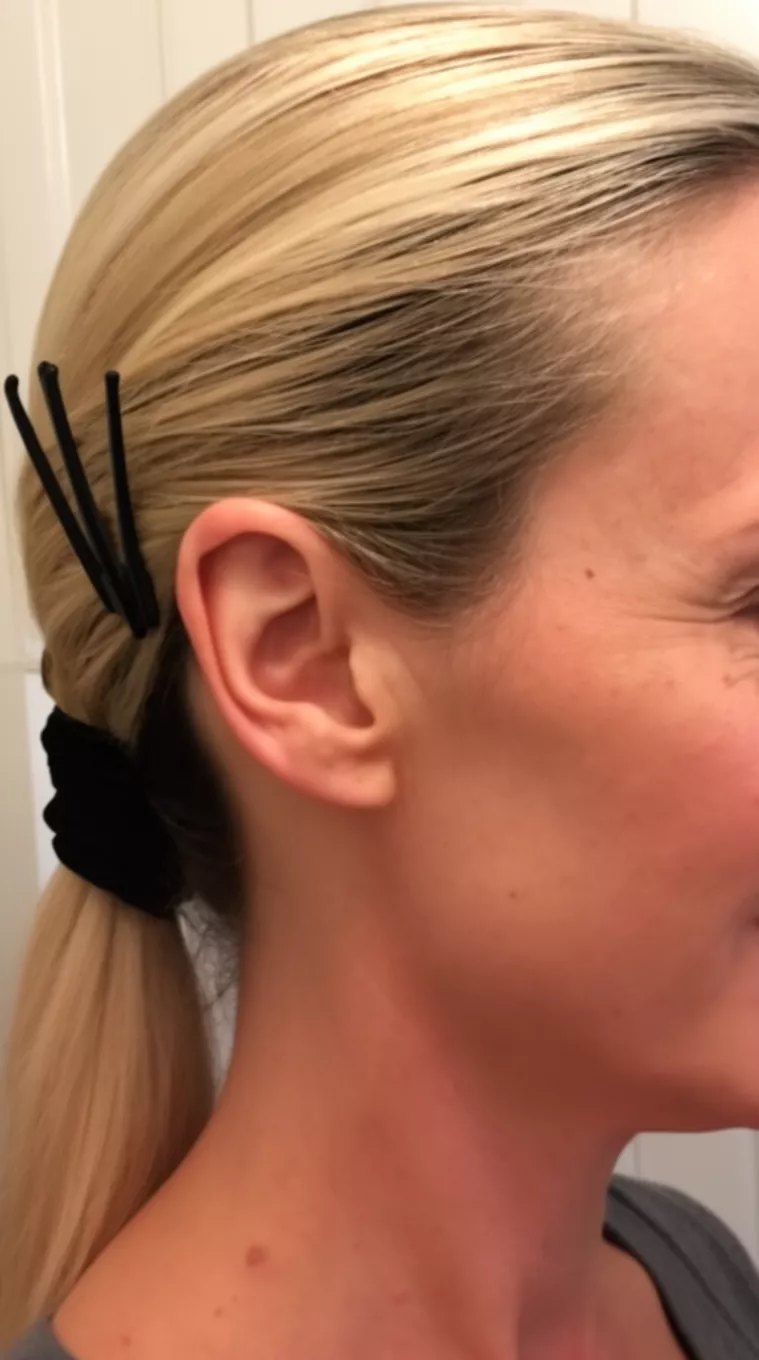 casual close-up side view photo of a 42-year-old woman with blonde hair pulled back slickly into a low ponytail secured with a black wrap and black barrettes near the ear, minimal background