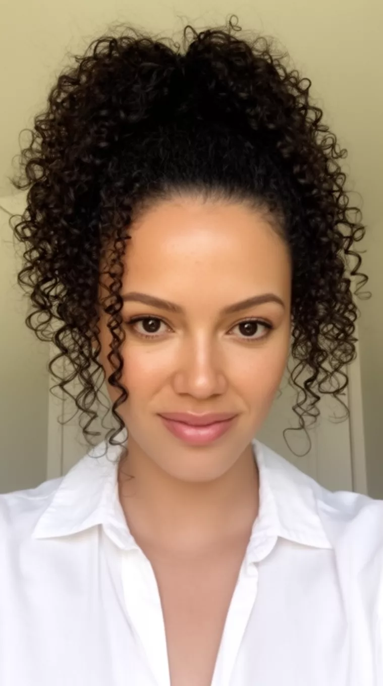 casual close-up photo of a 36-year-old woman wearing a white collared top with dark voluminous curly hair in a high elaborate bouncy ponytail, minimal background
