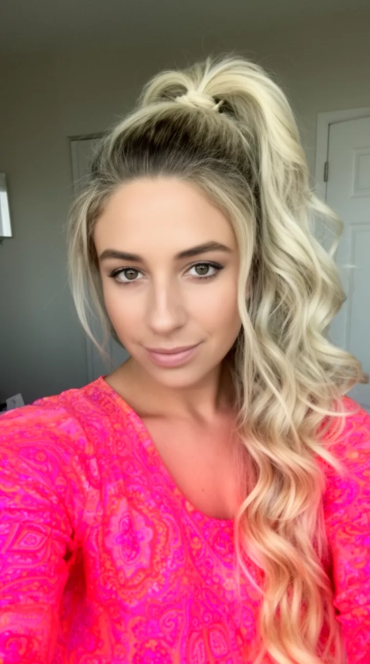 selfie of a 31-year-old woman wearing a patterned pink and orange top with blonde hair in a very high voluminous ponytail with long loose waves, minimal background