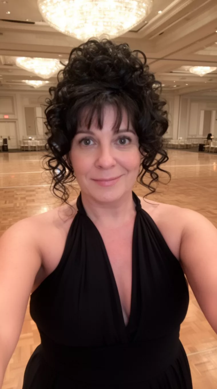 selfie of a 44-year-old woman wearing a black halter-neck dress with a dark hair colored curly updo with face-framing tendrils hairstyle, front view, empty ballroom background.