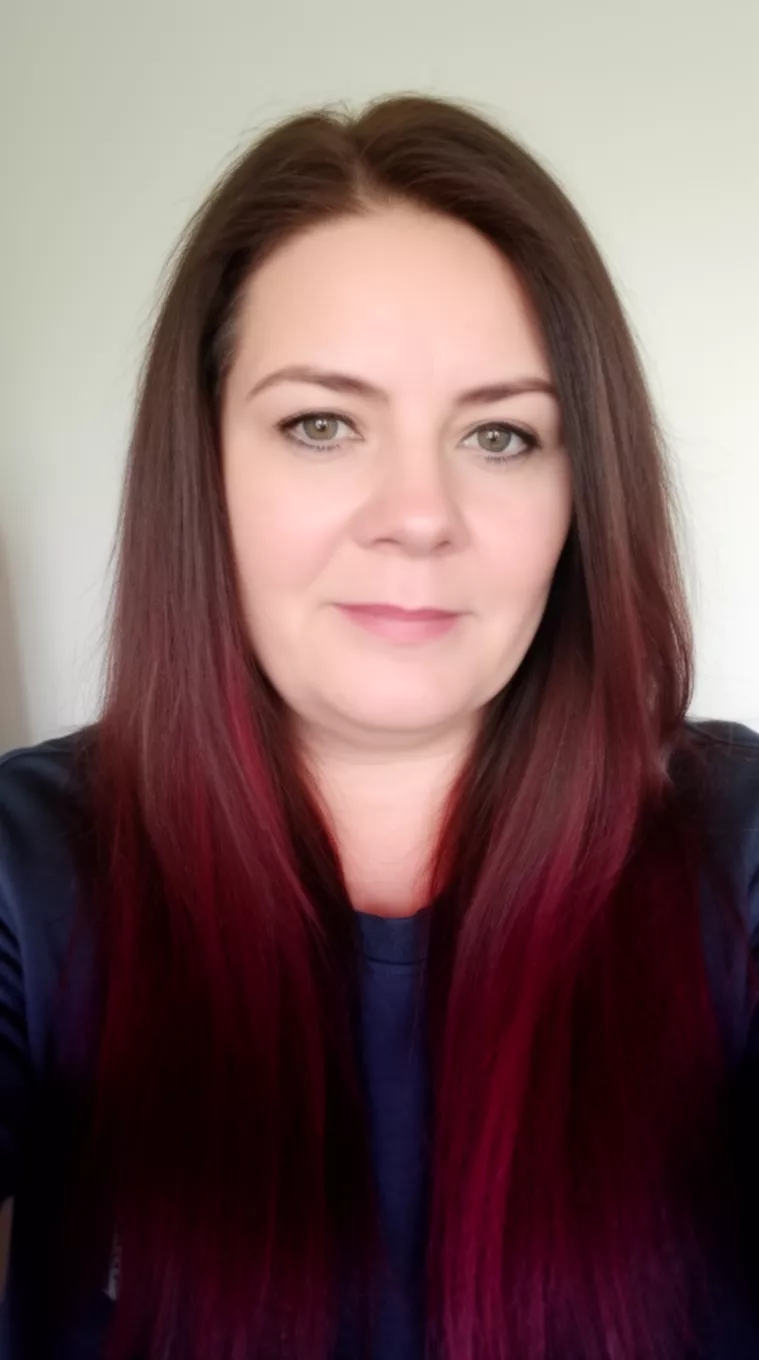 selfie of a 44-year-old woman wearing a dark top with a dark brown with burgundy highlights colored long hairstyle, front view, minimal background.