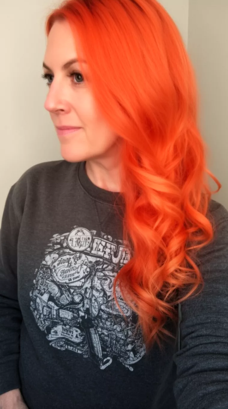 selfie of a 42-year-old woman wearing a dark grey graphic sweatshirt with a bright orange-red colored long wavy hairstyle, side view, minimal background.