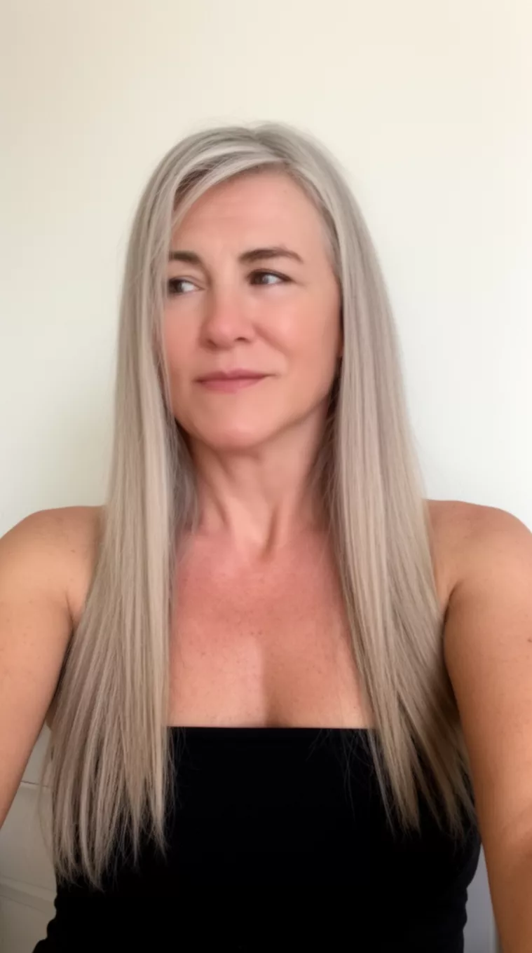 Subtle Layered Ash Blonde selfie of a 44-year-old woman wearing a black strapless top with an ash blonde colored long straight hairstyle, side view, minimal background.