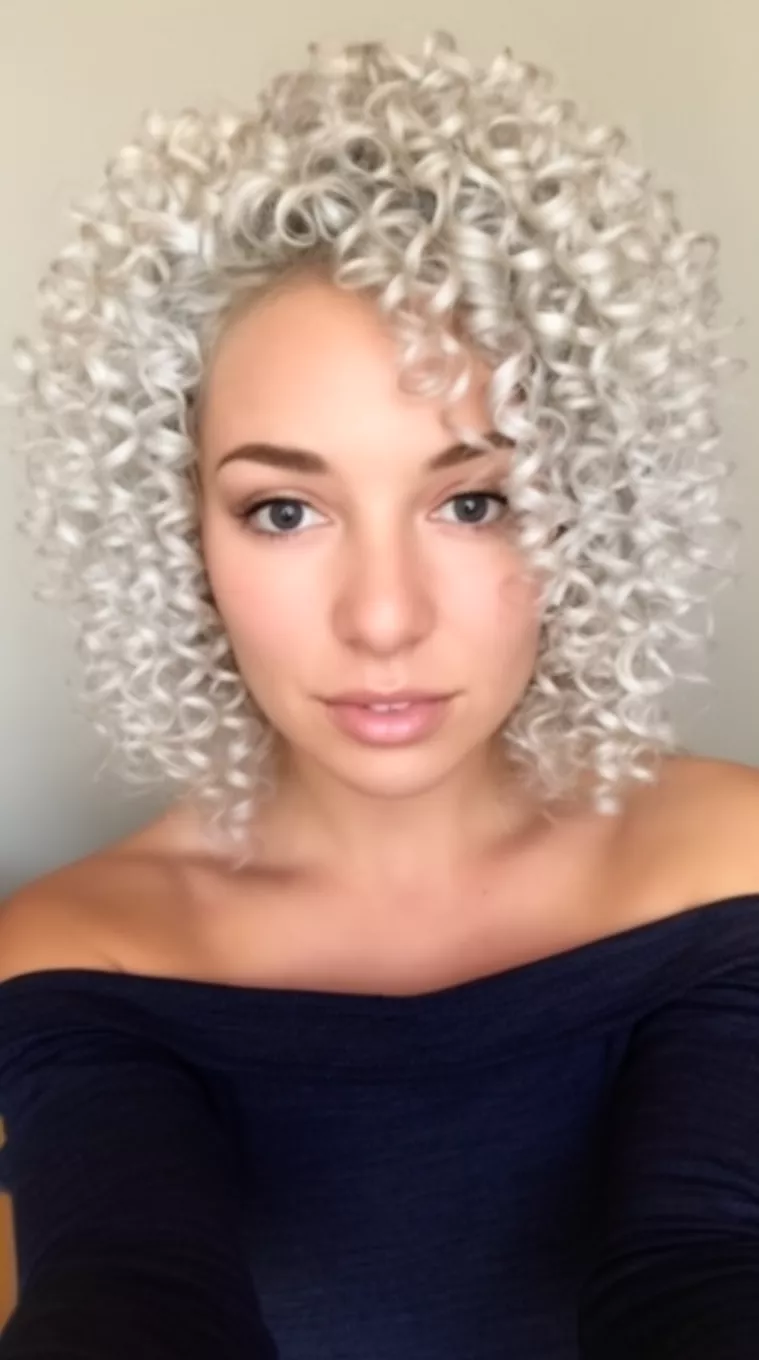selfie of a 29-year-old woman wearing a dark off-the-shoulder top with a Icy Platinum colored voluminous, platinum blonde, tight curls hairstyle, front view, minimal background.