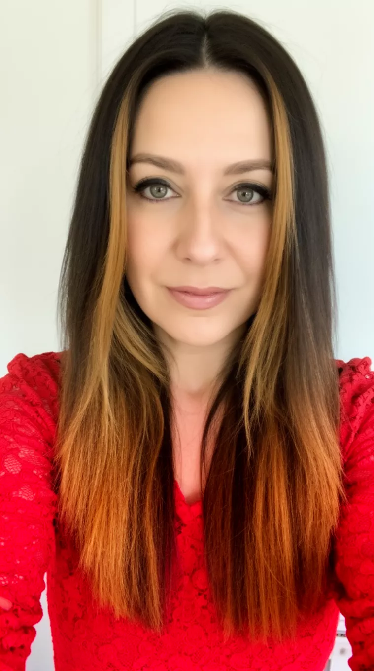selfie of a 39-year-old woman wearing a red lace top with a long deep brown with warm caramel highlights framing her face colored hairstyle, front view, minimal background.