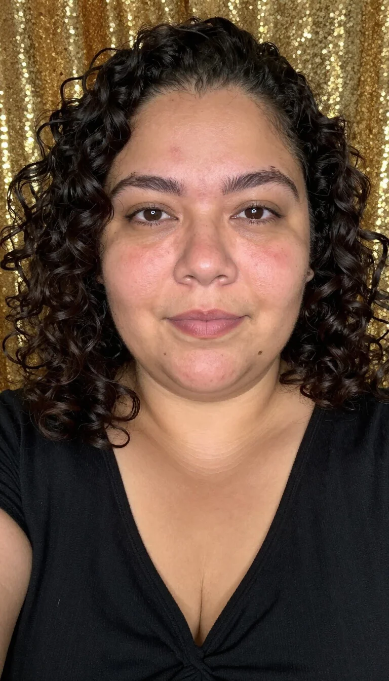 selfie of a 38-year-old plus size Latina woman with a chubby face and rounded nose. The skin is heavily textured with a complex pattern of fine lines around the eyes and a faint, thin scar traversing the left eyebrow. The eyes are dark brown. She is wearing a black knotted V-neck top with a dense dark curly hairstyle, front view, gold sequined background.