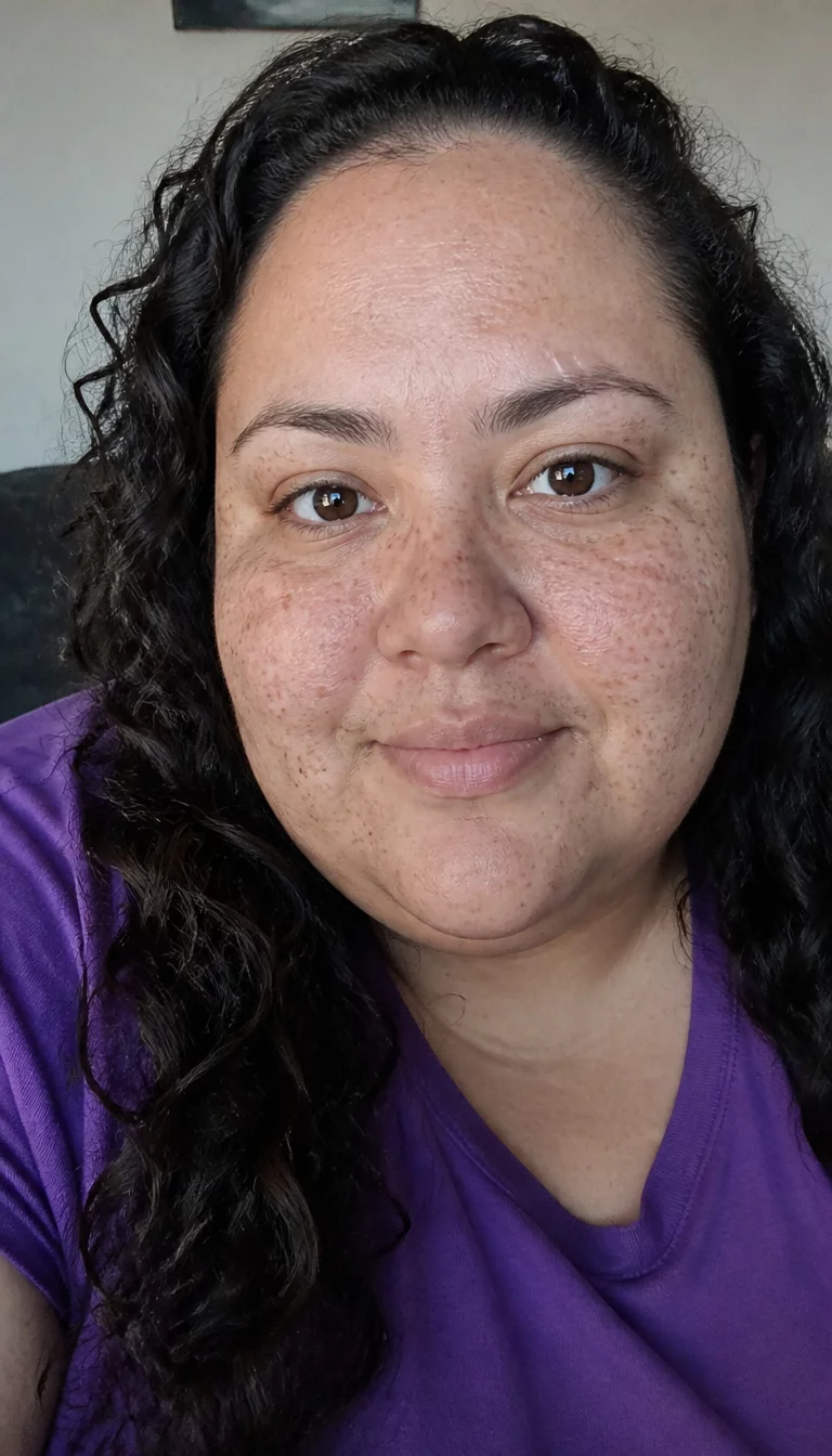 Freckles and Purple Tones selfie of a 36-year-old plus size Latina woman with a chubby face and delicate nose. The skin is heavily textured with a complex pattern of dark freckles and a faint, thin scar traversing the left eyebrow. The eyes are brown. She is wearing a purple shirt with a dark curly hairstyle, front view, natural lighting.
