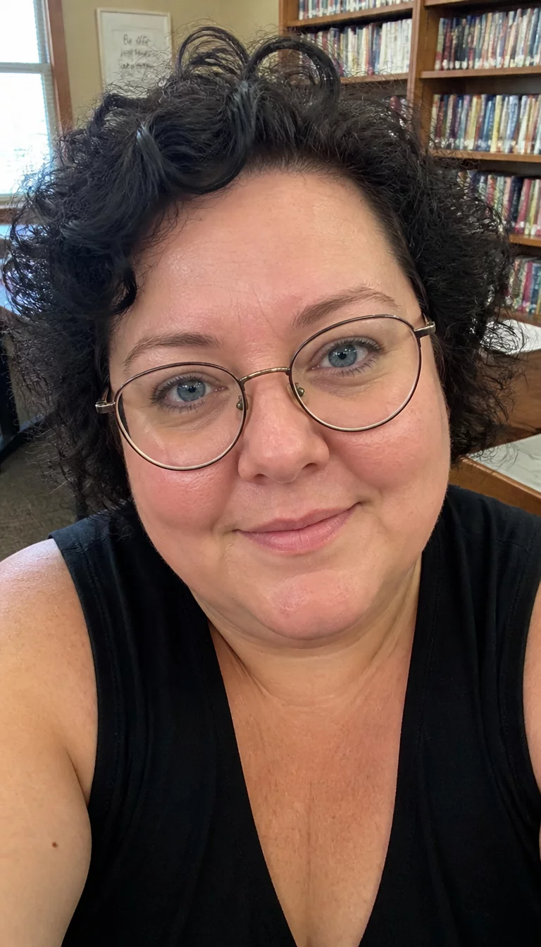 Academic Chic Short Curls selfie of a 41-year-old plus size Caucasian woman with a chubby face and delicate nose. The skin is heavily textured with a complex pattern of fine dry lines and a faint, thin scar traversing the left eyebrow. The eyes are blue. She is wearing a black sleeveless top and round glasses with a short dark curly hairstyle, front view, indoor library background.
