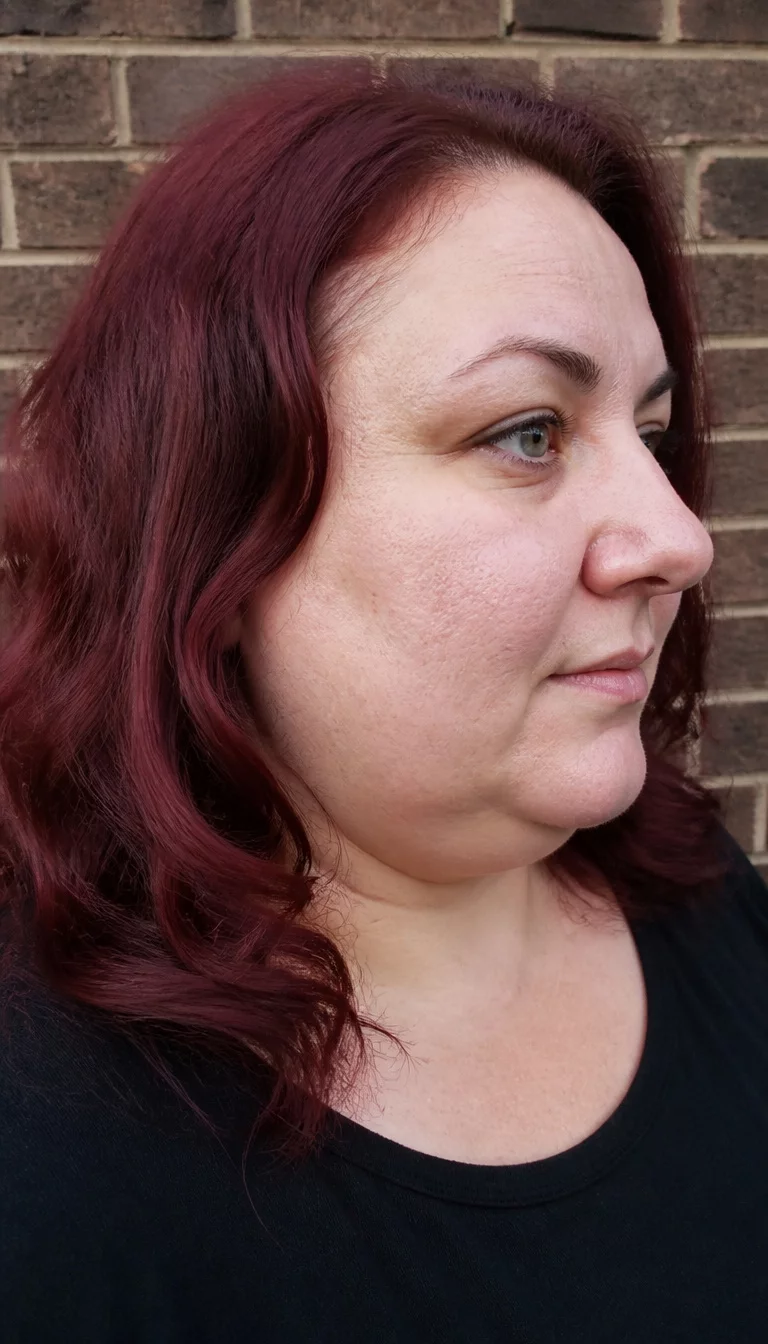 Modern Cherry Red Waves casual photo of a 42-year-old plus size Caucasian woman with a chubby face and pointed nose. The skin is heavily textured with a complex pattern of enlarged pores and a faint, thin scar traversing the left eyebrow. The eyes are gray. She is wearing a black top with a dark cherry red wavy shoulder-length hairstyle, side view, brick background.