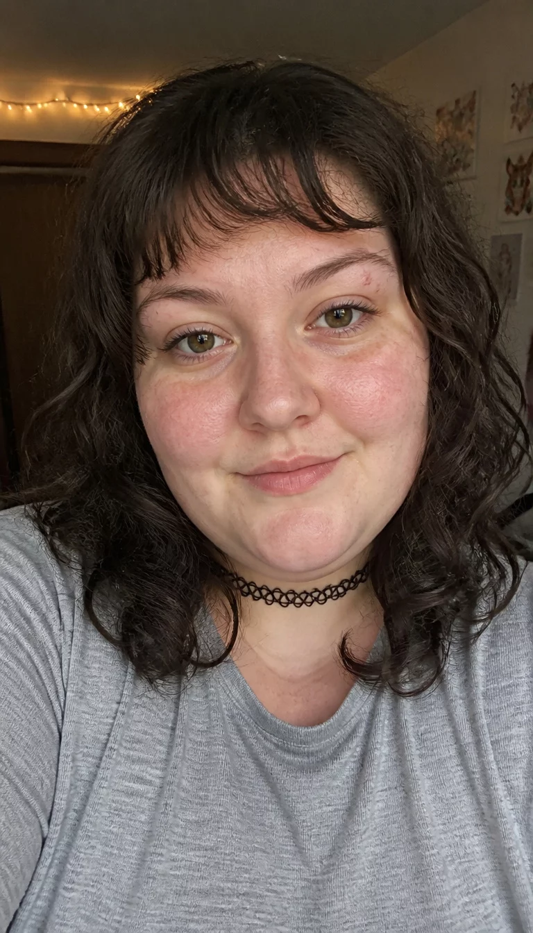 Edgy Curls with Bold Fringe selfie of a 27-year-old plus size Caucasian woman with a chubby face and delicate nose. The skin is heavily textured with a complex pattern of faint redness and a faint, thin scar traversing the left eyebrow. The eyes are hazel. She is wearing a grey shirt and black choker with a medium-length curly brown hairstyle and bangs, front view, indoor background.