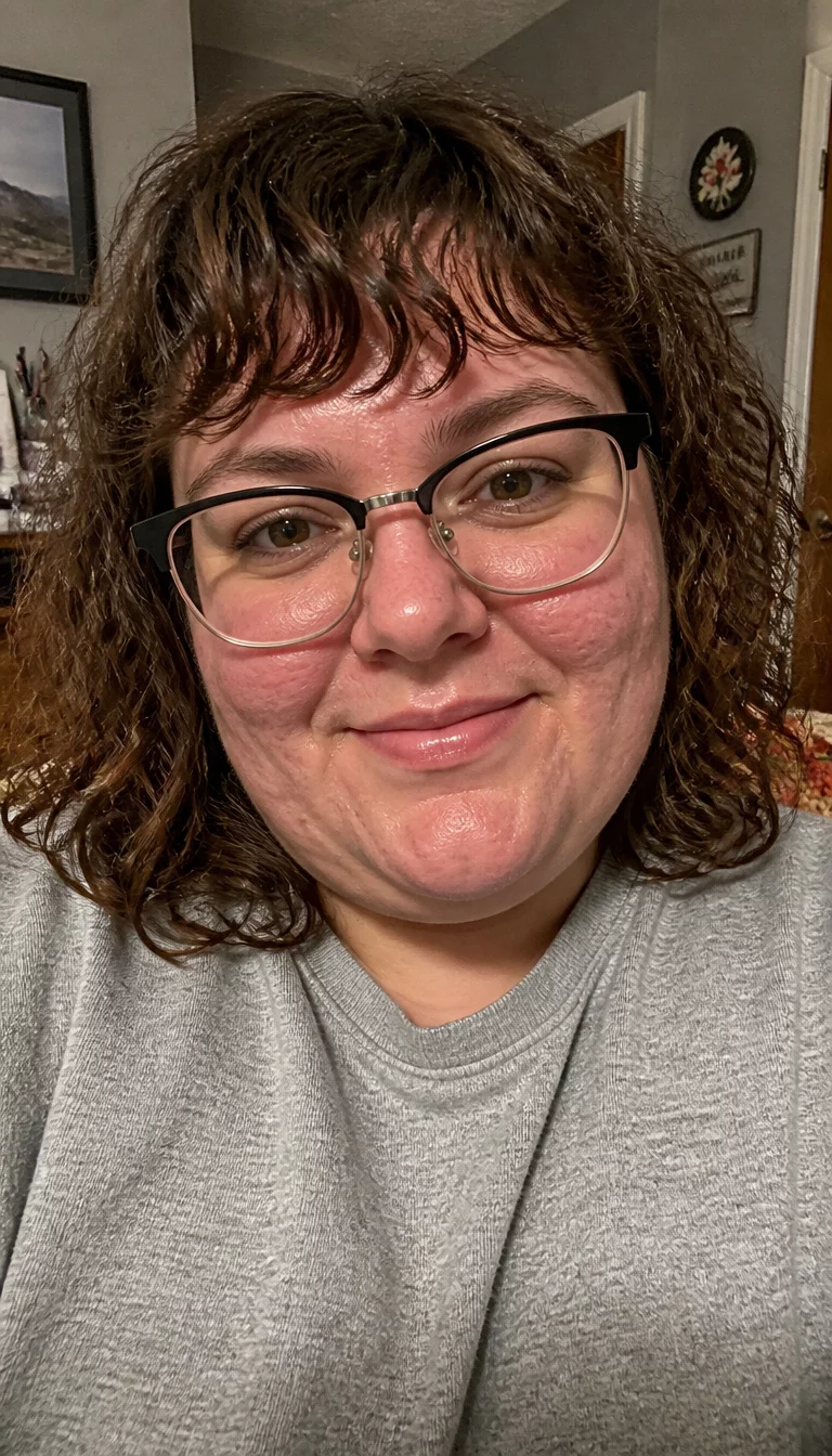 Casual Brown Curls with Bangs selfie of a 33-year-old plus size Caucasian woman with a chubby face and button nose. The skin is heavily textured with a complex pattern of acne scarring and a faint, thin scar traversing the left eyebrow. The eyes are light brown. She is wearing a gray sweatshirt and glasses with a shoulder-length curly brown hairstyle with bangs, front view, indoor home background.
