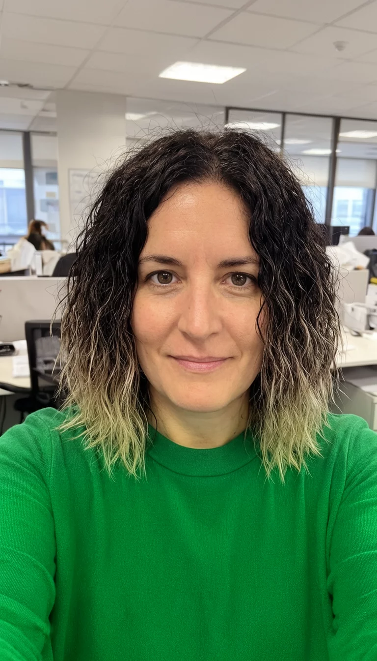 selfie of a 36-year-old woman wearing a green top with a dark origins and lighter ends colored textured lob hairstyle, front view, indoor office background
