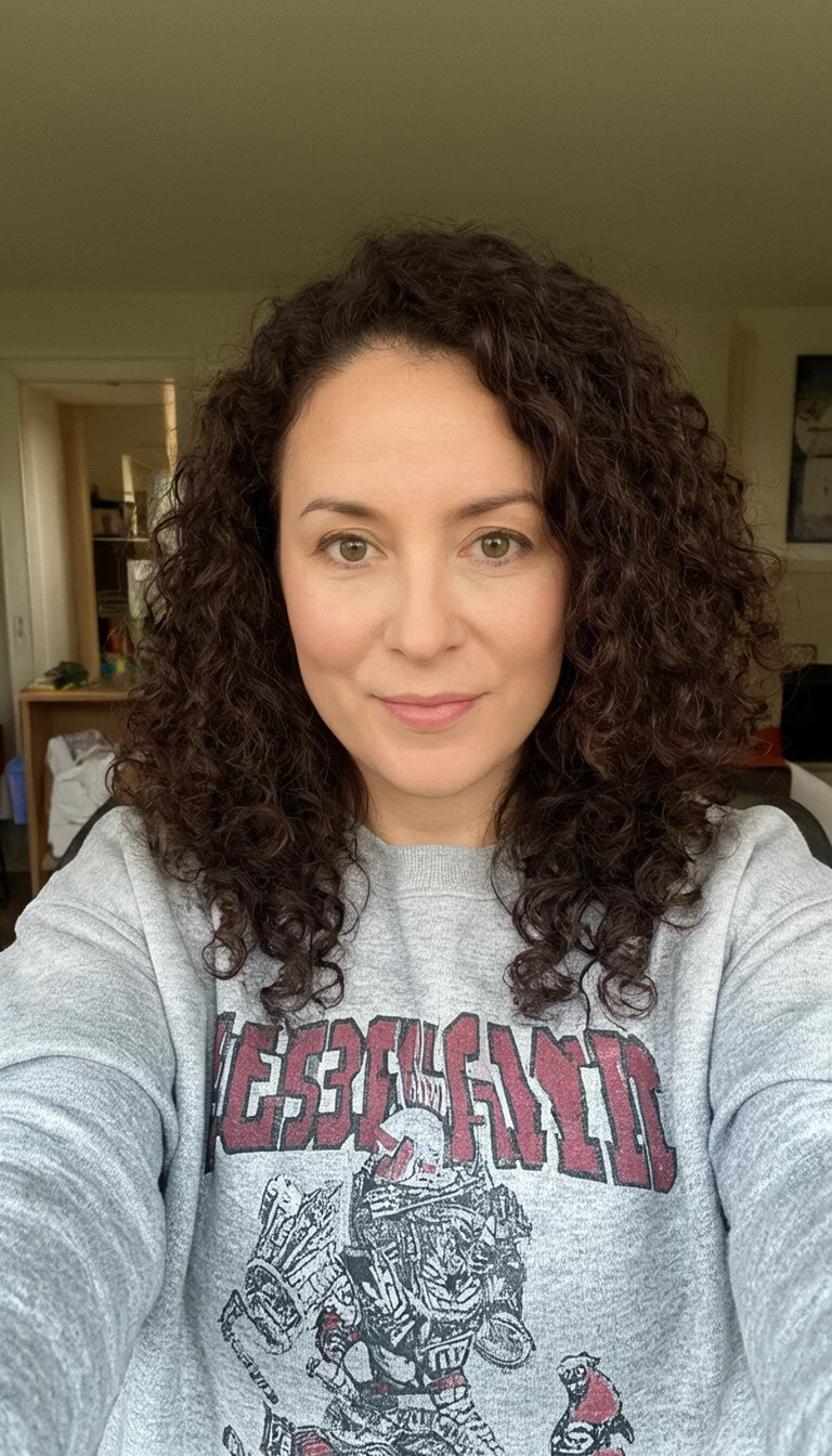 Classic Natural Deep Ebony selfie of a 34-year-old woman wearing a gray graphic sweatshirt with a dark brown colored curly hairstyle, front view, casual home background