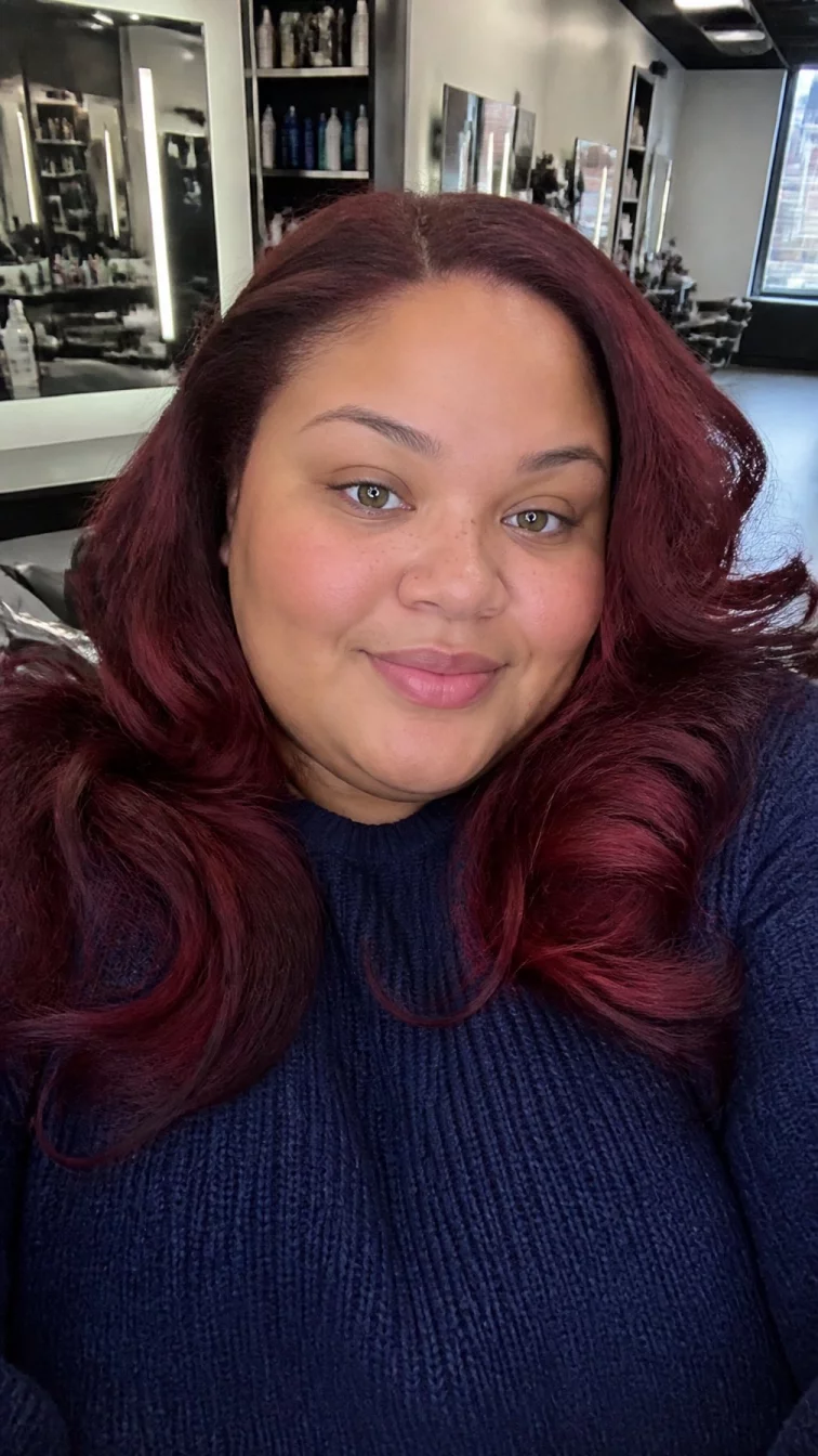 selfie of a 25-year-old Afro-Caribbean plus size woman with a chubby face, olive, upturned nose, straight eyebrows, and thin lips. Her face features faint freckles across the nose, slightly flushed cheeks, and subtle natural facial asymmetry. She has deep-set hazel eyes, rounded cheeks, closed-mouth smile, cheerful. She is average and is wearing a navy blue sweater with deep velvet burgundy voluminous waves hair, overcast daylight, hair salon