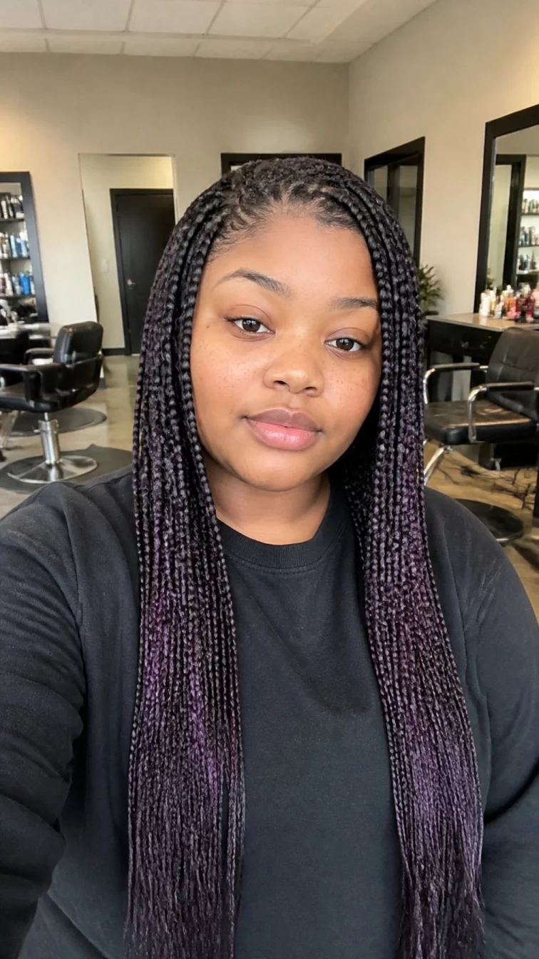 selfie of a 23-year-old Black African plus size woman with a chubby face, light, straight nose, softly curved eyebrows, and full lips. Her face features uneven eyebrows, light freckles, and subtle natural facial asymmetry. She has downturned hazel eyes, high cheekbones, neutral expression, relaxed. She is athletic and is wearing a black sweater with jet black hair with blackberry purple highlights braided hair, bright natural daylight, hair salon