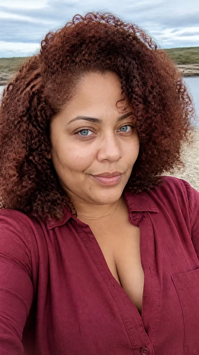 selfie of a 37-year-old Afro-Caribbean plus size woman with a chubby face, olive, aquiline nose, straight eyebrows, and full lips. Her face features faint freckles across the nose, faint under-eye shadows, and subtle natural facial asymmetry. She has hooded blue eyes, soft cheekbones, closed-mouth smile, playful. She is plus-size and is wearing a burgundy linen shirt with deep spiced auburn curly hair, overcast daylight, outdoor