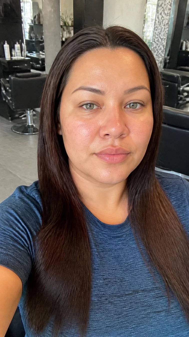 selfie of a 34-year-old Latina plus size woman with a chubby face, dark brown, broad nose, straight eyebrows, and full lips. Her face features widow's peak hairline, subtle acne marks, and subtle natural facial asymmetry. She has hooded green eyes, pronounced cheekbones, neutral expression, relaxed. She is slim and is wearing a denim blue athletic performance shirt with dark mahogany brown sleek straight hair, warm indoor lighting, hair salon