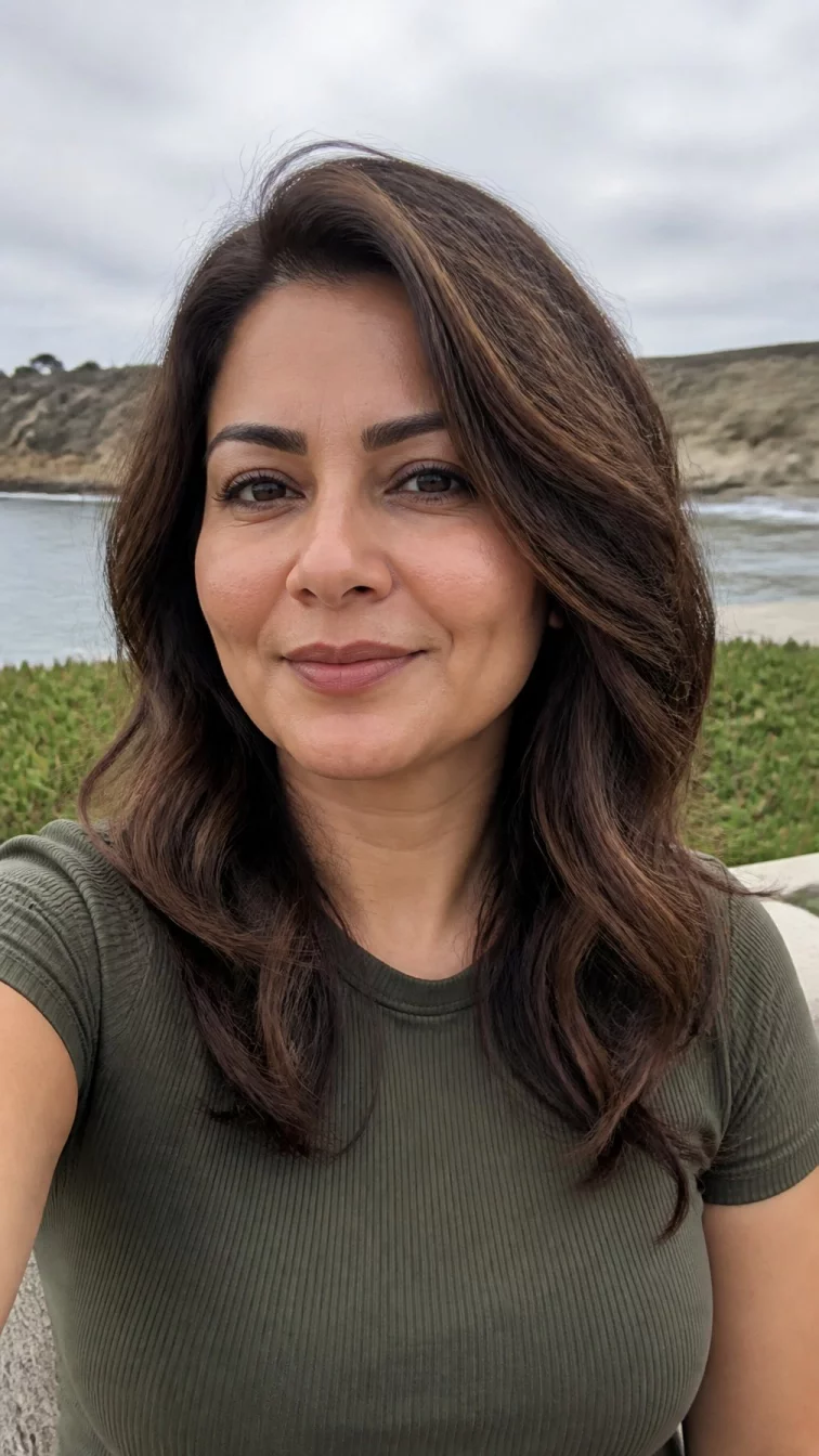 selfie of a 40-year-old South Asian woman with a square face, olive, upturned nose, feathered eyebrows, and heart-shaped lips. Her face features dimples, subtle acne marks, and subtle natural facial asymmetry. She has hooded dark brown eyes, soft cheekbones, closed-mouth smile, confident. She is petite and is wearing a olive green ribbed t-shirt with cinnamon swirl layered wavy hair, overcast daylight, outdoor