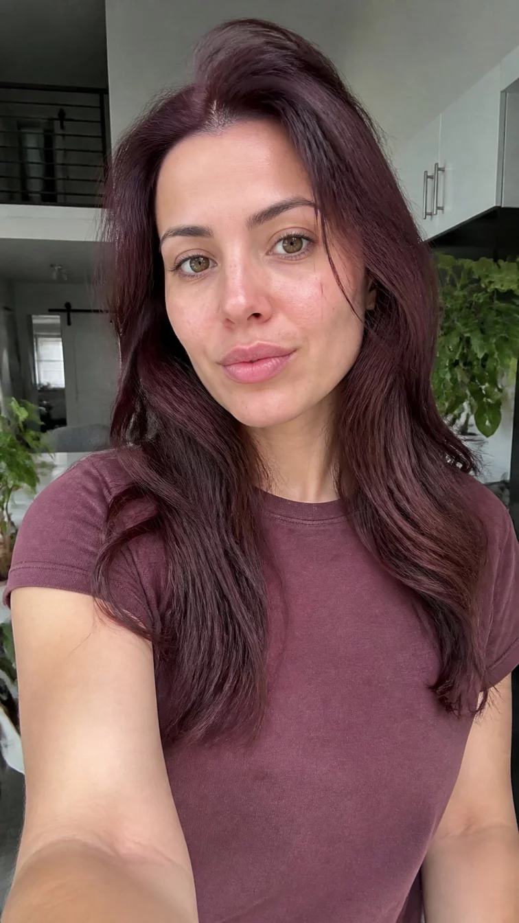 selfie of a 29-year-old Northern European woman with a diamond face, golden brown, upturned nose, feathered eyebrows, and wide lips. Her face features uneven eyebrows, a few laugh lines, and subtle natural facial asymmetry. She has almond-shaped hazel eyes, soft cheekbones, neutral expression, thoughtful. She is petite and is wearing a burgundy basic crew neck t-shirt with mulled wine burgundy waist-length wavy hair, bright natural daylight, indoor