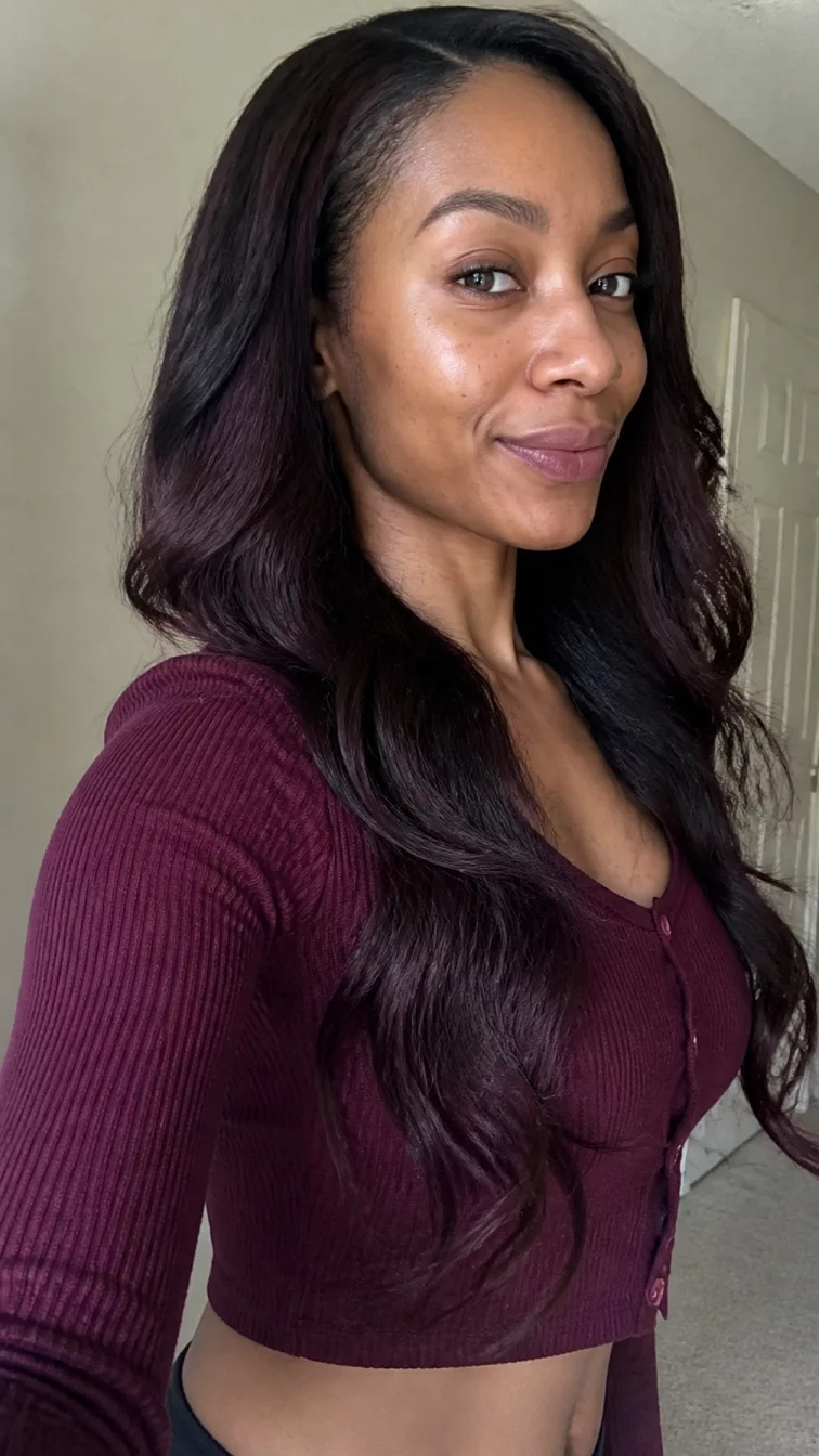 selfie A side photo of a 37-year-old Afro-Caribbean woman with a oblong face, dark brown, aquiline nose, thick natural eyebrows, and full lips. Her face features dimples, visible pores, and subtle natural facial asymmetry. She has deep-set gray eyes, rounded cheeks, closed-mouth smile, confident. She is slim and is wearing a burgundy cropped cardigan with plum charcoal long wavy hair, bright natural daylight, indoor