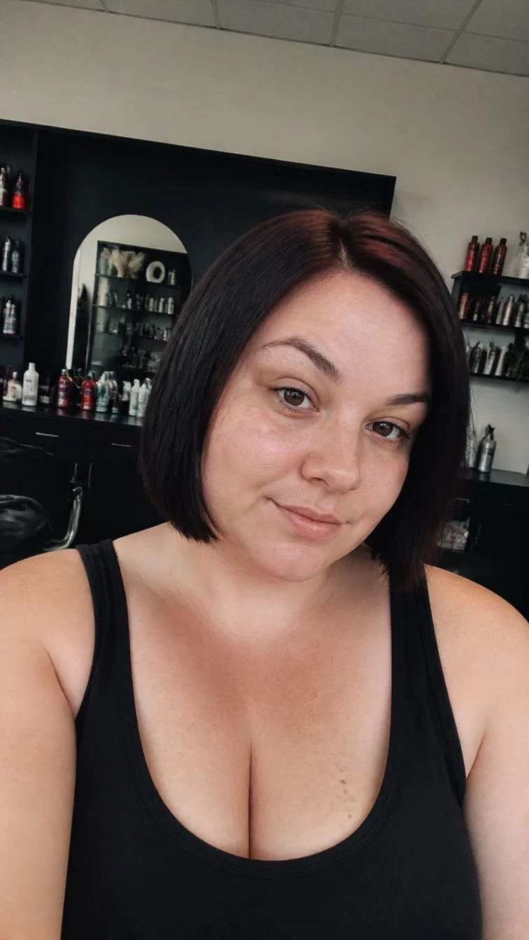 selfie of a 33-year-old Eastern European woman with a heart-shaped face, light, upturned nose, feathered eyebrows, and thin lips. Her face features small scar on the eyebrow, light freckles, and subtle natural facial asymmetry. She has deep-set dark brown eyes, rounded cheeks, neutral expression, playful. She is plus-size and is wearing a black tank top with glossy black cherry blunt bob hair, overcast daylight, hair salon
