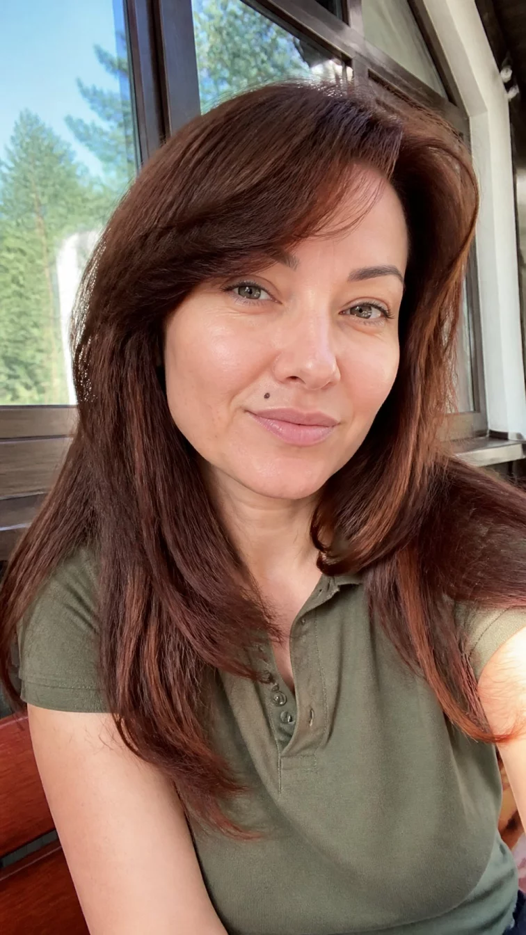 selfie of a 39-year-old Eastern European woman with a round face, dark brown, broad nose, feathered eyebrows, and thin lips. Her face features beauty mark near the lip, subtle acne marks, and subtle natural facial asymmetry. She has monolid gray eyes, soft cheekbones, closed-mouth smile, playful. She is slim and is wearing a olive green tennis polo with rich mahogany wood layered long hair, soft window light, outdoor