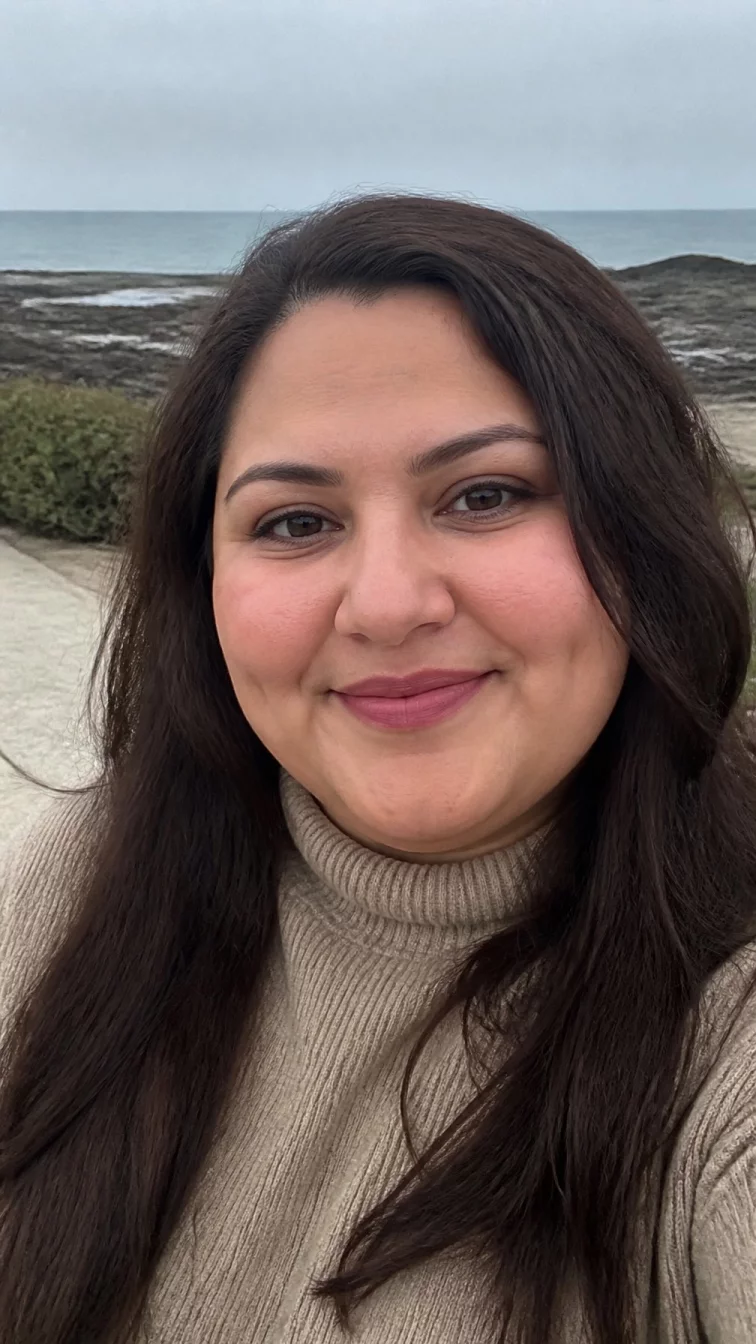 selfie of a 39-year-old Middle Eastern woman with a round face, light, rounded nose, feathered eyebrows, and full lips. Her face features dimples, subtle acne marks, and subtle natural facial asymmetry. She has round brown eyes, soft cheekbones, closed-mouth smile, cheerful. She is plus-size and is wearing a beige turtleneck sweater with deep cocoa bean long layers hair, overcast daylight, outdoor