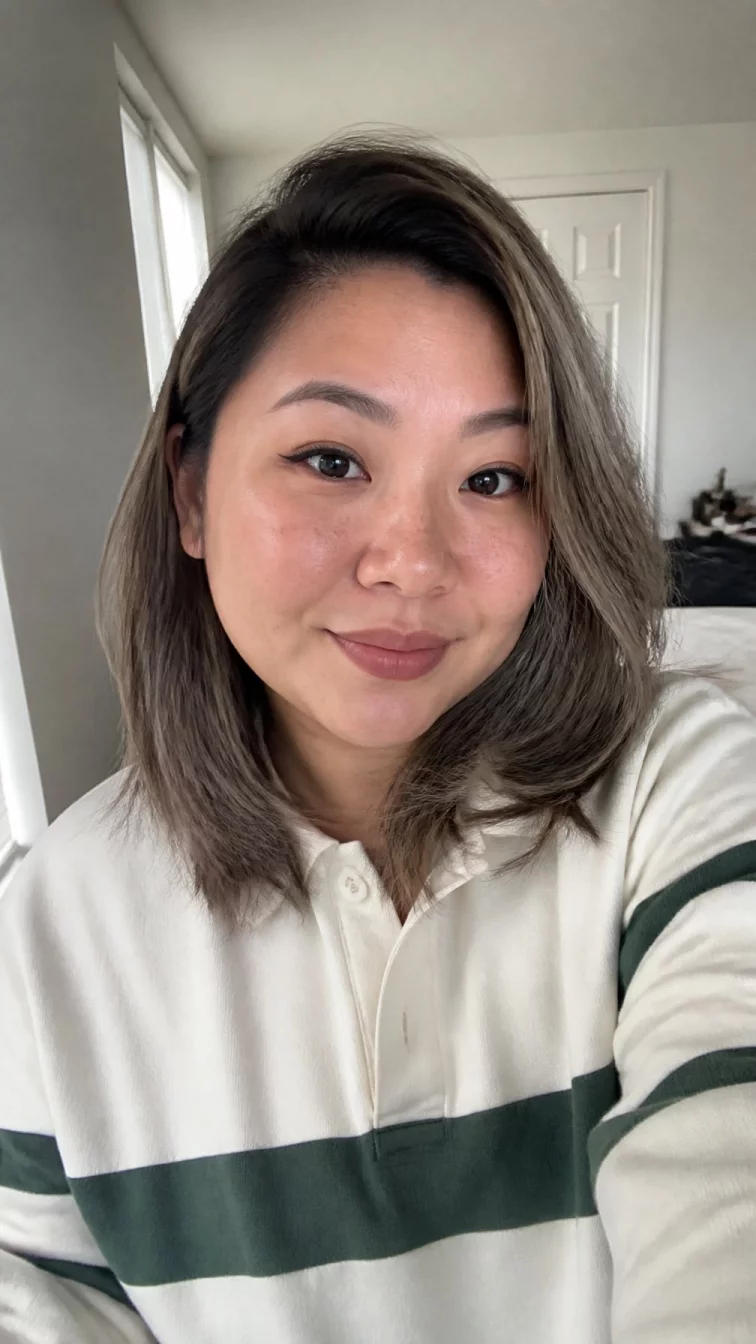 selfie of a 36-year-old East Asian woman with a heart-shaped face, dark brown, upturned nose, straight eyebrows, and heart-shaped lips. Her face features slightly crooked smile, light freckles, and subtle natural facial asymmetry. She has upturned gray eyes, soft cheekbones, subtle smile, playful. She is plus-size and is wearing a cream rugby shirt with silver sage ash brown lob hair, soft window light, indoor