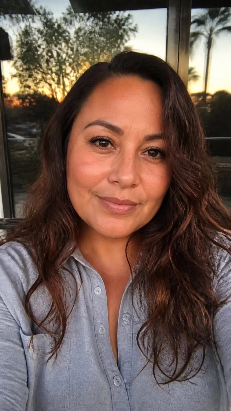 selfie of a 41-year-old Latina woman with a round face, tan, straight nose, thick natural eyebrows, and thin lips. Her face features slightly crooked smile, a few laugh lines, and subtle natural facial asymmetry. She has deep-set amber eyes, pronounced cheekbones, subtle smile, thoughtful. She is plus-size and is wearing a light gray button-up shirt with amber ale brunette wavy hair, warm indoor lighting, outdoor