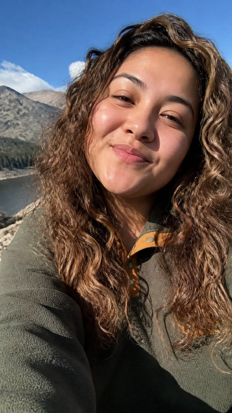 selfie of a 29-year-old Latina woman with a square face, fair, broad nose, softly curved eyebrows, and full lips. Her face features slightly crooked smile, subtle acne marks, and subtle natural facial asymmetry. She has monolid brown eyes, soft cheekbones, broad smile, playful. She is athletic and is wearing a olive green fleece pullover with golden brown with spicy nutmeg highlights voluminous curls, bright natural daylight, outdoor