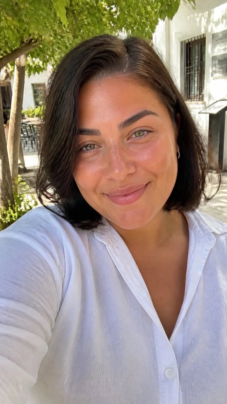 selfie of a 29-year-old Southern European woman with a heart-shaped face, golden brown, upturned nose, feathered eyebrows, and full lips. Her face features slightly crooked smile, slightly flushed cheeks, and subtle natural facial asymmetry. She has monolid green eyes, high cheekbones, closed-mouth smile, relaxed. She is plus-size and is wearing a white chambray shirt with dark chocolate brown with deep espresso lowlights sleek bob, bright natural daylight, outdoor