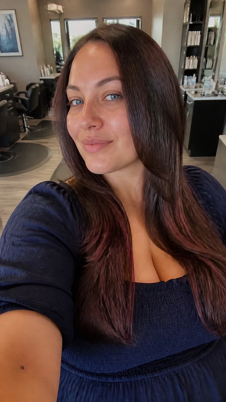 selfie A back photo of a 34-year-old Latina woman with a oval face, light, narrow nose, softly curved eyebrows, and full lips. Her face features faint freckles across the nose, light freckles, and subtle natural facial asymmetry. She has hooded blue eyes, high cheekbones, subtle smile, playful. She is plus-size and is wearing a navy blue smocked blouse with espresso brown with muted purple-brown highlights long straight hair, overcast daylight, hair salon