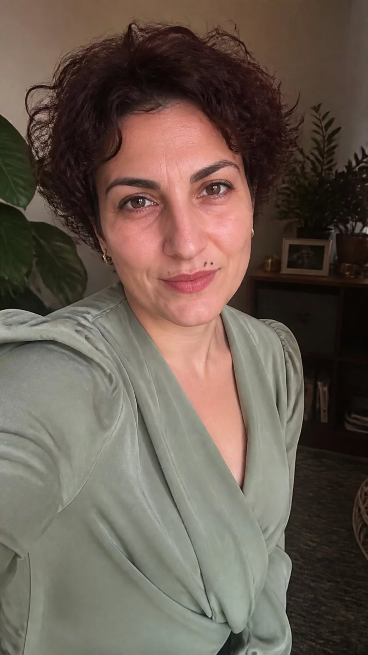 selfie of a 30-year-old Mediterranean woman with a round face, golden brown, aquiline nose, thick natural eyebrows, and thin lips. Her face features beauty mark near the lip, a few laugh lines, and subtle natural facial asymmetry. She has deep-set dark brown eyes, soft cheekbones, subtle smile, thoughtful. She is petite and is wearing a sage green wrap blouse with rich mahogany wine curly pixie hair, warm indoor lighting, indoor