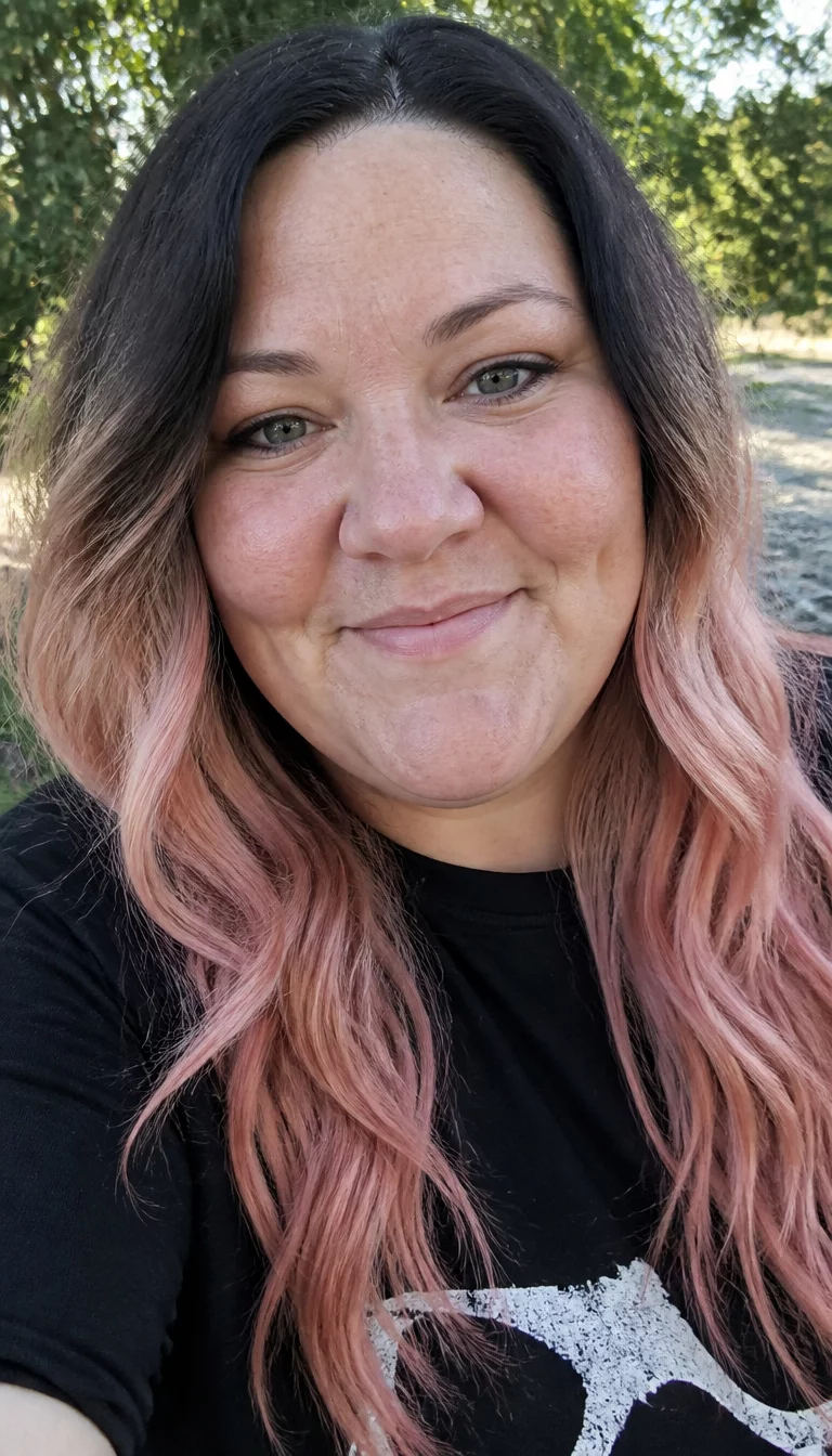 Dark To Pinkish Blonde Transition selfie of a 38-year-old plus size Caucasian woman with a chubby face and a slightly wide nose. The skin is heavily textured with a complex pattern of uneven pigmentation and a small dimple on the right cheek. The eyes are hooded grey. She is wearing a dark t-shirt with long, wavy hair transitioning from dark roots to pinkish-blonde ends, front view, outdoor background.