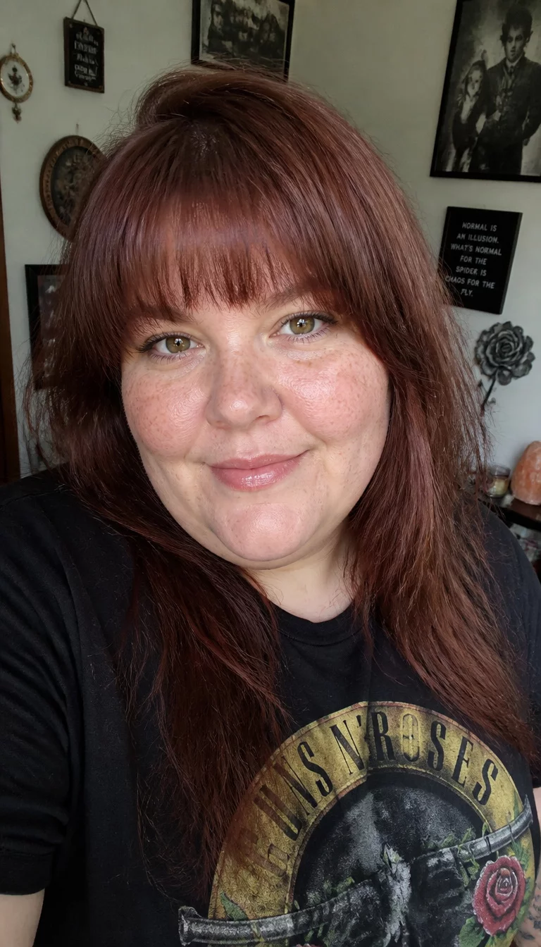 selfie of a 34-year-old plus size Caucasian woman with a chubby face and a straight, rounded nose. The skin is heavily textured with a complex pattern of light sun spots across the cheeks and a small beauty mark near the chin. The eyes are almond shaped hazel. She is wearing a black graphic t-shirt with a reddish-brown, layered hairstyle with full bangs, front view, room with decorated walls.
