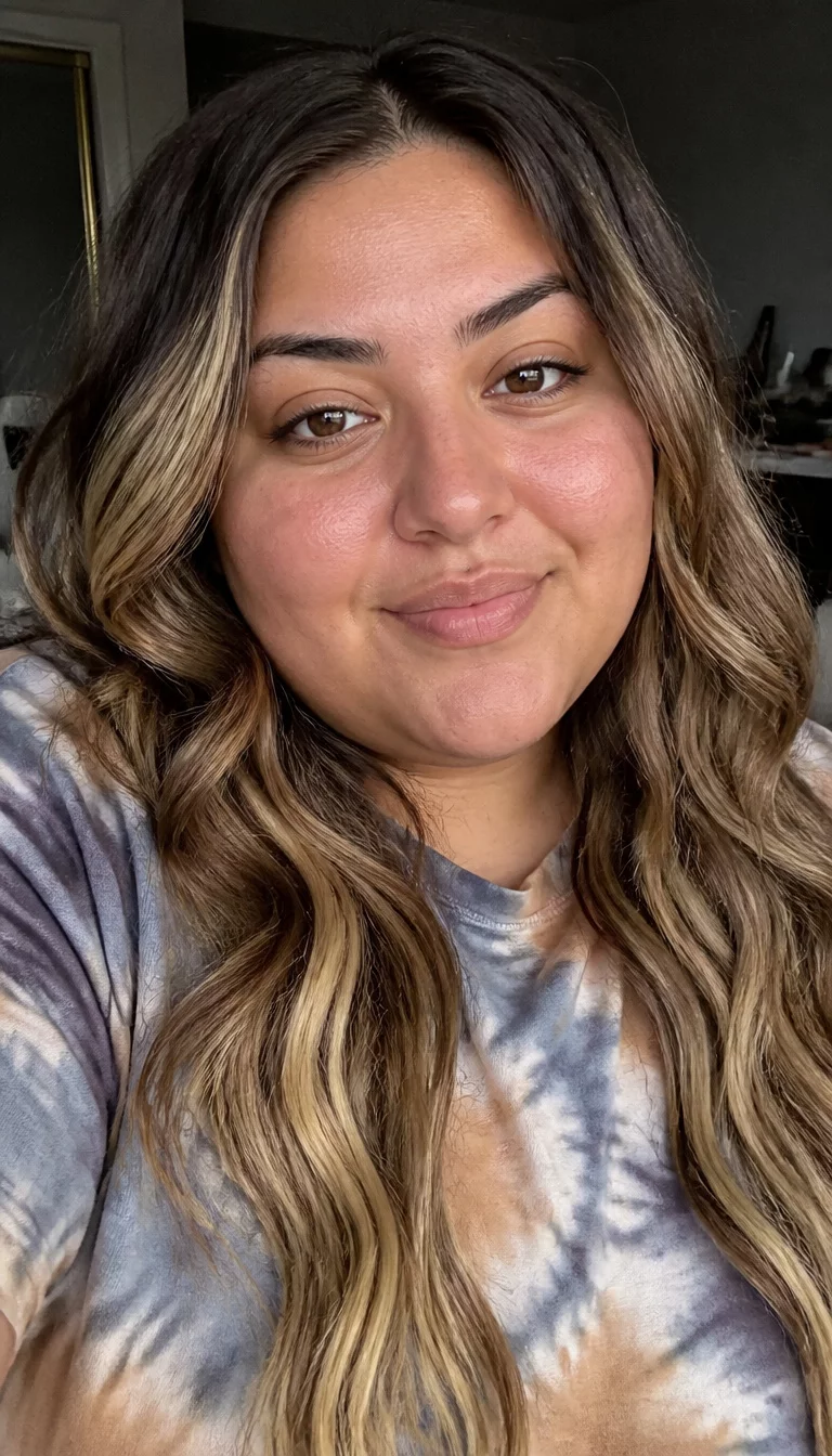 selfie of a 29-year-old plus size Middle Eastern woman with a chubby face and a slightly flared nose. The skin is heavily textured with a complex pattern of olive undertones and a tiny freckle on the upper lip. The eyes are almond-shaped dark brown. She is wearing a tie-dye t-shirt with a dark brown and blonde balayage hair styled in loose waves hairstyle, front view, indoor setting.
